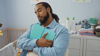 Sakallı genç adam rahat bir yatak odasında kitap tutuyor. İçinde bebek malzemeleri, oyuncaklar ve bebek malzemeleri var. Huzurlu bir ev ortamını yansıtıyor..