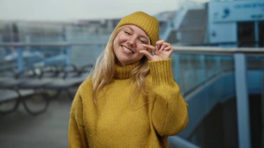 Sarı kazaklı kadın güvertede gülümsüyor. Küçük bir el hareketi yapıyor. Açık deniz ortamında neşeli bir hava sergiliyor..