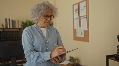 Çalışma ortamında odak ve profesyonelliği yansıtan çizelge ve kitaplarla dolu bir ofis içinde, panoya yazı yazan yaşlı bir kadın..