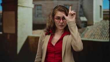 Gözlük takan bir kadın, eski bir üniversitenin iç mekanında, tarihi mimariye karşı kırmızı gömleğini ve bej ceketini vurguluyor..