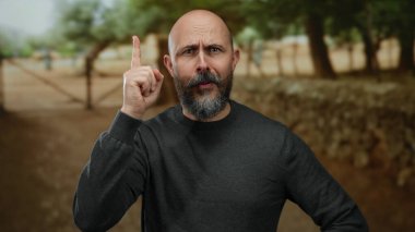 Kel sakallı bir adam, etrafı bulanık ağaçlarla çevrili bir açık hava parkında düşünceli bir şekilde el kol hareketi yaparak doğal ve sakin bir ortamda tefekküre dalmış..