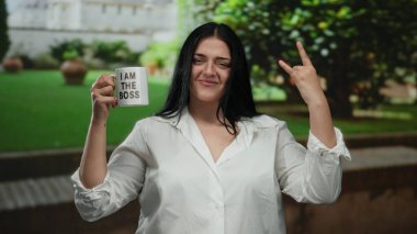 Siyah saçlı, beyaz tişörtlü, patron kupası tutan genç bir kadın yeşil park ortamında kaya jesti yapıyor. Açık havada güven ve güç gösterisi yapıyor..