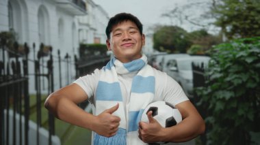 Sokakta gülümseyen ve elinde başparmağıyla futbol topu tutan atkı takan genç bir adam açık hava sporları coşkusu ve şehir yaşam tarzını öneriyor..