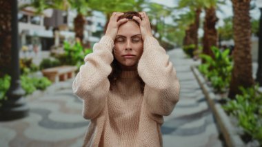 Kazaklı kadın, açık hava parkında hayal kırıklığını ifade ediyor. Palmiye ağaçları ve bankların bulanık manzarasıyla..