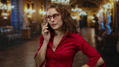 Kırmızı tişörtlü bir kadın kilisenin içinde avizelerle süslü telefonlar kullanıyor. Etrafı loş ışıklandırma ve zarif dekorlarla çevrili. Sohbete odaklanmış görünüyor..