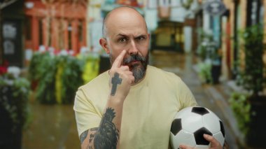 Kentsel caddede futbol topu tutan kel adam meraklı bir şekilde gözünü dikip dövmesini gösteriyor ve günlük sarı gömleğini giyiyor..