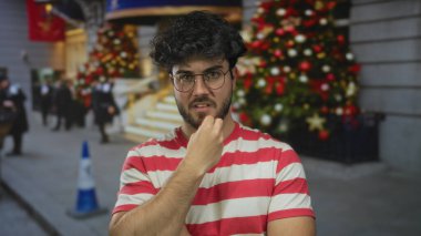 Sakallı ve gözlüklü genç adam arka planda Noel süsleri ve ağaçlarla dolu bir sokakta geziniyor..