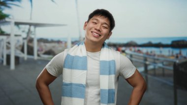 Çizgili atkısı olan genç bir adam sakin bir okyanus arkaplanı olan hareketli bir sahil sahilinde gülümsüyor açık havada rahat ve kaygısız bir yaz havasını somutlaştırıyor..