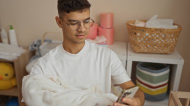 Genç İspanyol bir adam hastane odasında bebeği tutuyor, telefona bakıyor, klinik ortamında yeni doğan bebekler için şefkatli ve besleyici bir ortam öneriyor..