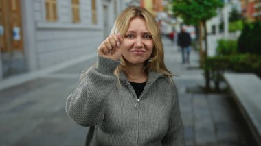 Şehir caddesinde süveter giyen bir kadın iletişim kurmayı öneriyor, arka planda binalar olan açık şehir ortamında duygularını aktarıyor..