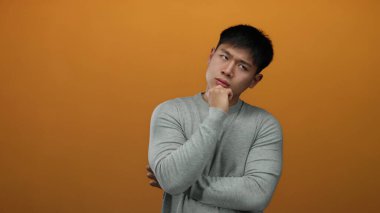 Genç adam, izole edilmiş turuncu bir duvara karşı düşünceli bir ifadeyle düşünüyor. Düşünce ve yansıma anı sergiliyor..