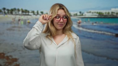 Sahilde neşeli hareketler yapan sarışın bir kadın. Canlı bir ifadeyle. Arka planda sakin bir plaj manzarası var..