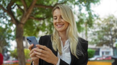Şehir parkında akıllı telefon kullanan, gülümseyen sarışın şehir ortamından hoşlanan, teknolojinin ve doğal ortamdaki boş zamanların özünü yakalayan bir kadın..