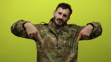 Askeri üniformalı İspanyol sakallı bir adam, kendine güvenen, kendine güvenen, kendine güvenen ve odaklanan sarı bir arkaplanı işaret ediyor..