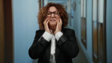 Otel koridorunda stresli hisseden bir kadın, orta yaşlı özelliklerini vurgulayan gözlükler, iş kıyafetleri giymiş, kapalı bir ortamda duygu ve endişeyi ifade eden bir kadın..
