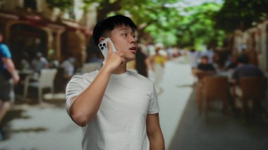 Açık kafe terasında telefonla konuşan genç adam, beyaz gömlek giyiyor, canlı yeşillik ve hareketli restoran atmosferiyle çevrili..