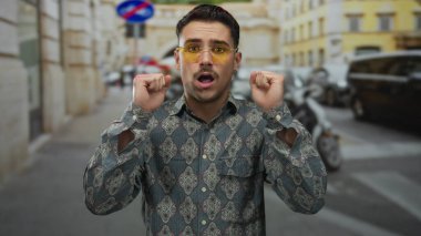 Şaşkın bakışlı İspanyol genç adam renkli binalar ve park edilmiş arabalarla şehir merkezinde şık gözlükler takıyor..