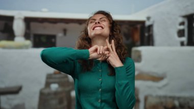 Sokak ortamında yeşil süveter giyen, şehir yaşamını ve canlı ifadeyi yansıtan beyaz arka planda gülümseyen hashtag jesti gösteren bir kadın..