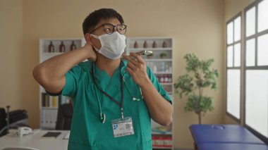 Genç doktor ameliyat önlüğü ve maske takarak tıbbi ekipman ve doğal ışıkla çevrili modern bir klinikte akıllı telefonu kullanarak düşünceli ifadesini geliştiriyor..