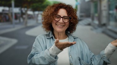 Orta yaşlı İspanyol bir kadın, bir şehir caddesine uzanmış gözlüklü ve kot gömlekli neşeli bir ifade sergiliyor..