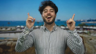 Arka planda gemilerle limanda gülümseyen sakallı genç İspanyol adam çizgili bir gömlek giyerek güneşli bir günün tadını çıkarıyor ve yüzünde deniz esintisini hissediyor.