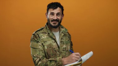 Askeri üniformalı İspanyol sakallı adam not defterine canlı turuncu arka plan hakkında yazıyor..