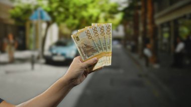 Bir şehir caddesinde Norveç kronu banknotları tutan adam, şehir ortamında açık hava finans kavramını vurguluyor, para ve yaşam tarzına odaklanmış bir şekilde yakalanıyor..