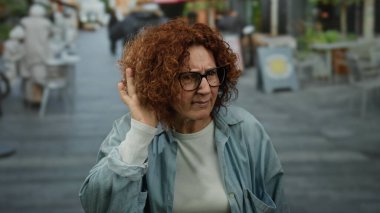 Orta yaşlı, kızıl saçlı ve gözlüklü bir kadın kalabalık bir şehir caddesinde merakla ve nişanlanmayla ilgili bir jest yapıyor..