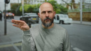 Gri kazaklı kel bir adam, arka planda gizemli bir atmosferi taşıyan araçlarla şehir caddesinde gizemli bir jest yapıyor..
