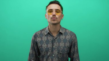Şablonlu gömlekli ve sarı gözlüklü genç adam canlı yeşil arka planda duruyor. Kendine güvenen ve şık görünüyor..