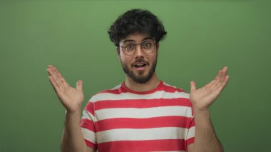 Çizgili tişörtlü, sakallı ve gözlüklü genç bir İspanyol stüdyo ortamında yeşil arka plana karşı şaşırmış görünüyor..