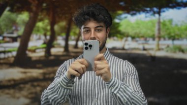 Sakallı, elinde araba anahtarı ve akıllı telefonuyla gölgeli yeşil bir parkta güneşli bir günün tadını çıkaran genç bir İspanyol..