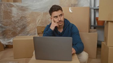 Etrafı kutularla çevrili yeni bir evdeki genç adam dizüstü bilgisayar kullanarak oturma odasında düşünce jesti yapıyor..