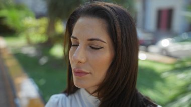 Şehir manzarasıyla çevrili şehir parkında meditasyon yapan bir kadın, gözleri kapalı, açık havada huzurlu bir anın tadını çıkarırken doğal ışıkla özelliklerini ön plana çıkarıyor..