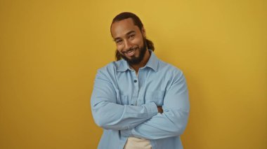 Sakallı, gülümseyen bir adam açık mavi gömlekli, parlak sarı arka plana karşı duruyor. Kollarını kavuşturmuş, kendine güvenini ve pozitifliğini ifade ediyor.