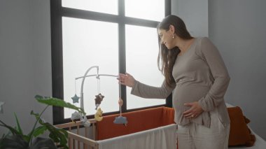 Yatak odasında hamile bir kadın büyük bir pencereden içeri süzülen yumuşak bir ışıkla çevrili bir beşiğin üstündeki cep telefonuna hafifçe dokunuyor..