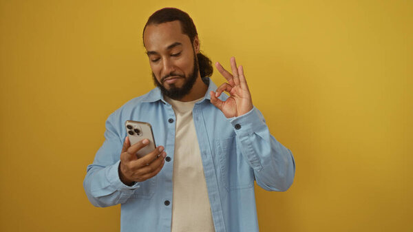 Man with beard holding smartphone against yellow wall making an ok gesture while wearing a blue shirt, looking content and relaxed in a casual setting.