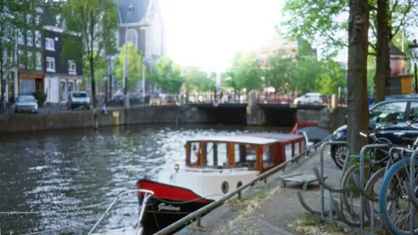 Güneşli bir günde yemyeşil ağaçların altındaki yürüyüş yolu boyunca rıhtımlı bir tekne ve bisikletlerle Amsterdam 'daki bir kanalın bulanık görüntüsü, Hollanda' nın kentsel manzarasının özünü yakalıyor..