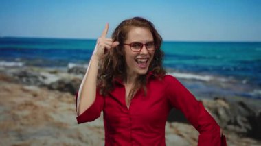 Kırmızı gömlekli kadın deniz kenarında neşeyle gülüyor, açık mavi deniz arka planına işaret ediyor, kumsaldaki mutluluk ve heyecanı somutlaştırıyor..