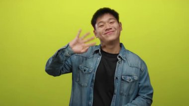 Sarı arka planda gülümseyen ve kot ceketli genç Çinli adam dostça bir selamlama ve olumlu bir ruh hali içinde.