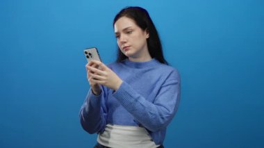 Esmer saçlı bir kadın mavi arka planda akıllı telefon kullanıyor, günlük mavi kazak giyiyor, ciddi bir ifadeyle ekrana bakıyor, kapalı alanda izole edilmiş..