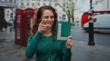 Şehirli bir çevrede seyahat eğlencesi ve açık hava deneyimini gösteren kırmızı telefon kulübesi olan öğrenci tabelasıyla gülümseyen bir kadın..