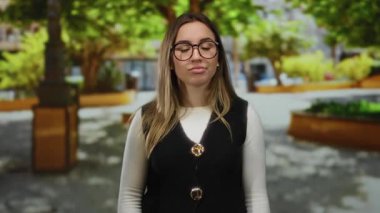 Gözlüklü genç bir kadın, dışarıda güneşli bir parkta yeşil ve ağaçlarla çevrili bir yelek ve beyaz gömlek giyerek düşünceli bir poz veriyor..