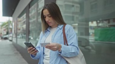 Şehir caddesinde elinde telefon ve kart tutan, günlük mavi gömlek giyen, bilgileri kontrol eden, cam bir binanın yanında duran, odaklanmış yüzlü, sarı saçlı bir kadın..