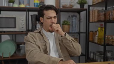 Ofis ortamında masada dinlenen genç adam, ofis malzemeleri ve mutfak malzemeleriyle çevrili, bej ceket giyiyor, yorgun ve çalışma saatlerinde uyanık kalmaya çalışıyor..