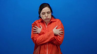 Turuncu tulumlu, esmer saçlı, mavi arka planda soğuk görünen, kapalı bir ortamda rahatsızlığını dile getiren bir kadın..