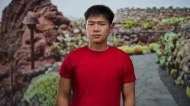 Kırmızı tişörtlü genç Çinli adam kayalık bir yolda dışarıda dikilirken elleriyle gözlerini kapatıyor..