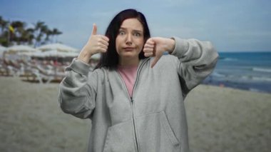 Güneşli bir sahil sahnesinde açık mavi deniz ve uzak palmiye ağaçlarıyla karışık duygular ifade eden bir kadın..