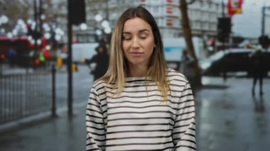 Sarı saçlı, çizgili tişörtlü genç bir kadın şehir caddesinde duruyor, bulanık şehir aktiviteleriyle çevrili, yağmurlu bir ortamda çeşitli duyguları ifade ediyor..