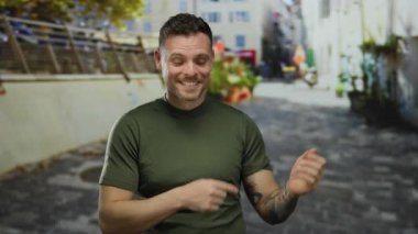 Yeşil gömlekli genç adam şehir caddesinde duruyor. Dövmesi görünür, canlı şehir zeminiyle çevrili, odaklanmış bir ifadeyle modern şehir yaşamını gözler önüne seriyor..